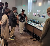 Image of an EPA scientist talking while standing in front of microscopes and agar plates surrounded by 5 people listening