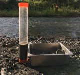 Image of a stream with a rocky shore. There is a metal container with silt sitting on the shore with next to a test beakers with a  water and sediment sample.