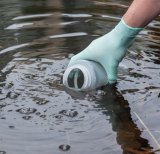 Person's hand with a light blue glove holding a water sample collection sample vial. They are dipping the vial into a body of water to collect a water sample.